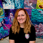 Jessica sits in front of several of her colorful paintings. She is wearing a black t-shirt and is smiling.