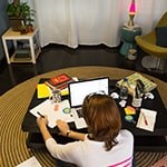 A student sits cross-legged on the floor in front of a coffee table with a laptop, papers, notebooks, and pens.