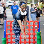 A student smiles while wearing headphones around their neck, sunglasses, a blue headband, and a dark shirt. A giant Connect 4 game and a crowd of people are behind them. The words “Student Community Association Throws Campus Life Block Party” are displayed on the photo.