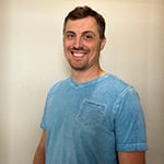 James Lane stands in front of a beige wall and smiles at the camera. He is wearing a light blue v-neck t-shirt.