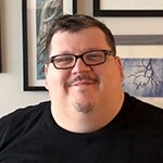 A smiling man wearing glasses and a black t-shirt sits in front of a wall covered with framed paintings.