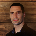 Justin Hosie standing in front of a paneled wood background. He is wearing a dark denim shirt and gently smiling.