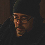 Michael Romano stands in front of a grey wall wearing a beanie, hoodie, and glasses with a pencil tucked behind his ear.