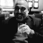 Omar Ortiz smiles as he holds a glass in his hand. He is wearing a dark jacket.