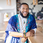 Rico Robinson stands in a café. He is wearing a blue-patterned wrap over a white button-up shirt. He is smiling.