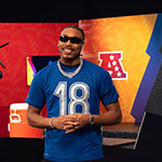 Minnesota Vikings wide receiver Justin Jefferson smiles while wearing a blue NFL-branded t-shirt with the number '18' in a studio set for the Madden NFL 25 Boom Challenge.