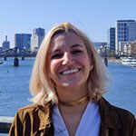 Grad Angela Decastro, a woman with shoulder-length blonde hair wearing a brown jacket and white top, smiles while standing on a bridge overlooking a river, in the distance behind her is a bridge and cityscape.