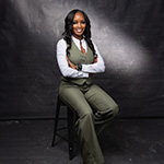 A woman in a green vest and pants sits confidently on a stool, arms crossed, against a dark studio backdrop.