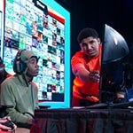 Two Florida high school students seated on stage in front of a gaming monitor with a Full Sail Armada athlete helping them to their right.