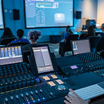 A person is seen operating a professional audio mixing console in a classroom, with students seated and screens displaying sound software.