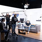 A group of students work together in a living room set on one of Full Sail’s soundstages. They are using a camera and holding microphones and a script.