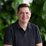 A man with short brown hair is wearing a dark grey polo while smiling against a lush green backdrop.