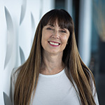 A woman with long brown hair and bangs smiling while standing against a geometric white backdrop.