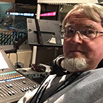 A man seated at an audio console overlooking a basketball court in a live arena.