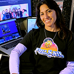 Instructor Milena Jackson, a woman with shoulder-length dark hair, is seated at her desk while wearing a Full Sail University graphic tee.