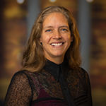 Phoebe Elefante, a woman with shoulder-length light brown hair and a black blouse smiling in front of an out-of-focus brown backdrop.