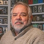 Randy sits in front of a wall covered by shelves of vintage cameras. He is wearing a light brown sweater.