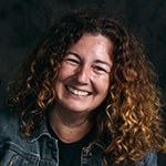 A woman with curly auburn hair smiling while wearing a black tee and denim jacket against a black backdrop.