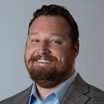 A man with short brown hair and a short brown beard smiles at the camera, wearing a plaid blazer and light blue shirt against a grey background.