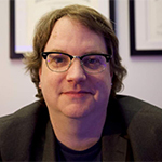 Up close headshot of a man wearing glasses and a black shirt against a wall with framed degrees.