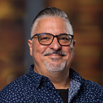 Alex Ginzo smiles at the camera. He is wearing a blue and white collared shirt and glasses, and he has gray hair and a short gray beard.