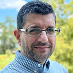 A man with short curly hair, glasses, and a trimmed beard smiles slightly while standing outdoors, the background is out of focus.