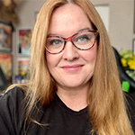 Amy Williams sits in her home design studio. The walls are covered in framed artwork. She is wearing glasses and smiling.