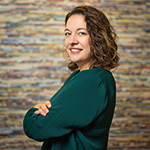 Aron Gelineau stands with her arms crossed and smiles at the camera. She has brown, curly hair and is wearing a teal sweater.