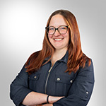 A smiling person with long red hair, glasses, and a blue zip-up jacket, is standing with their arms crossed against a light gradient background.