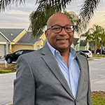 Chris stands outside wearing a grey blazer, blue shirt, and black-rimmed glasses. He is smiling.