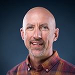 Corbett Compel smiles for the camera in front of a blue background. He is wearing a plaid shirt.