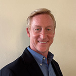 Greg Downey smiles at the camera in front of a light background. He is wearing a blue shirt and a dark blazer.
