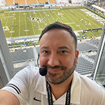 Jeff Sharon wears a headset microphone and smiles at the camera. He is in an announcer’s booth with the UCF football stadium below him.