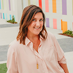 Jenn Miller sits in front of a colorful outdoor wall. She is smiling and wearing a pink shirt and a gold necklace.