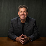 Joe Gornik sits at a wooden table and smiles at the camera. He is wearing a gray plaid jacket with a Full Sail employee pin on the lapel.