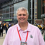A man with short white hair and a pink polo shirt is seen smiling while standing in front of Lambeau Field in Green Bay, Wisconsin.