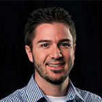 Matt is sitting in front of a black background wearing a blue plaid shirt. He is smiling.