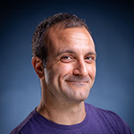 Michael Corinella smiles at the camera in front of a blue background. He is wearing a purple shirt.