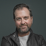 A man in a dark brown leather coat and light t-shirt appears from the waist up, smiling against a grey backdrop.