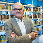 Rolando stands in a library with his arms crossed. He is smiling and wearing a suit.