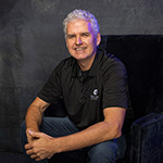 Roy Papp sits in a blue velvet chair in a photography studio. He is smiling and wearing jeans and a black polo shirt with a Full Sail logo.
