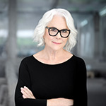 A woman with short white hair and glasses, wearing a black top, smiles confidently with arms crossed against a blurred background.