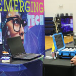 Two people at TechFest explore a driving simulator next to an ‘Emerging Tech’ display with gadgets and a laptop on the table.