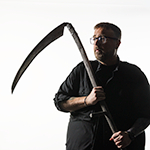 Full Sail Grad Aaron B. Koontz is seen wearing black as he holds a large scythe against a bright white background, gazing thoughtfully to the left side of the frame.