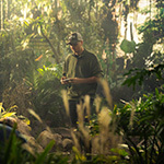 Nate Strayer stands on a film set filled with lush green trees and a digital forest background. He is wearing a baseball cap that says Stray Vista Studios.