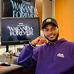 Alain sits at a desk with multiple monitors that read “Wakanda Forever” and “Marvel Studios.” He is wearing a purple hoodie.