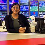 Priscilla sits at a desk in a live news television studio. She is wearing glasses and is smiling.