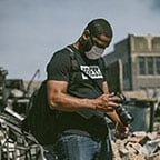 Grad E Mackey looking down at a DSLR camera while wearing a grey face mask, backpack, and black shirt with the word "press" in white, standing amongst a large amount of debris.