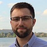 A man wearing glasses and a button-up shirt stands in front of a pier.