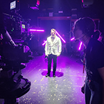 An actor stands in the middle of a set wearing a white suit and bowtie with purple lights around them. A student wears a headset and operates a camera pointed at the actor.
